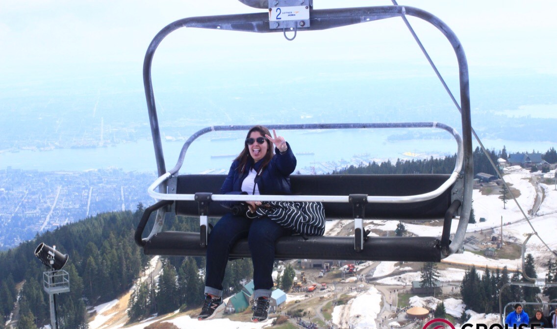 Chrys sentada no banco de um teleférico com a cidade de Vancouver ao fundo.